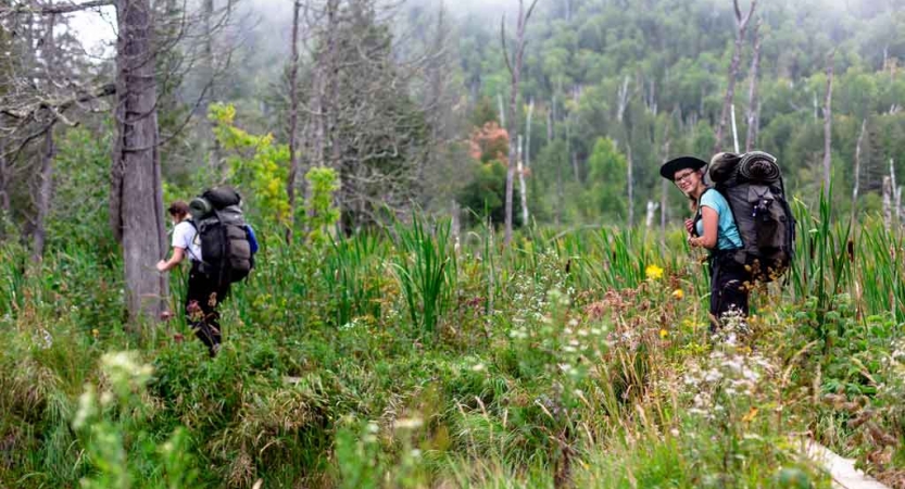 two outward bound students carrying backpacks hike through a green landscape